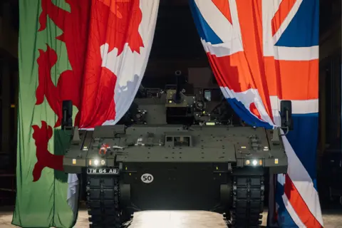 General Dynamics A green armoured vehicle is emerging between two large flags either side - a Welsh dragon on the left and a union flag on the right.