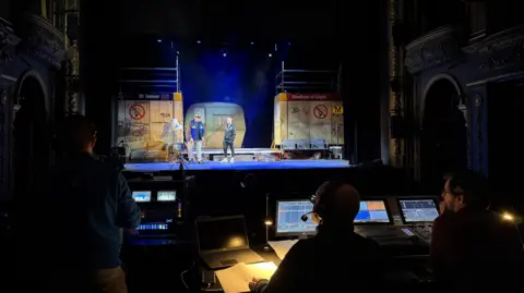 Production technicians sitting behind monitors while two actors on stage practice their performance. The set has been designed to look like a Metro station, with St James and one side and Stadium of Light at the other.