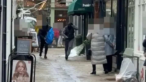 Shot of two men wearing black balaclavas and black and grey clothing running down a narrow cobblestone street. Two women onlookers are blurred out and are looking towards them.