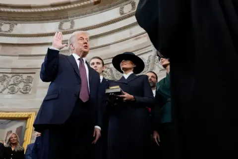 Morry Gash/AP Donald Trump, wearing a dark suit and purple tie, stands with his right hand raised as Melania Trump, in a dark suit and dark hat, holds the Bible during the 60th Presidential Inauguration.  The curve of the Rotunda ceiling is in the background 