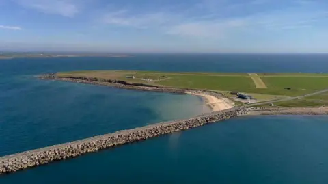 An aerial view of the Churchill Barriers at Scapa Flow - a large expanse of water with a causeway linked to a flat green island.