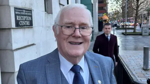 A close-up image of Eugene Reavey. He is an older man with medium-length white hair. He is wearing a pair of metal framed glasses and a grey checked suit jacket and waistcoat. He is also wearing a white shirt and a blue patterned tie. He is standing outside the Belfast High Court.