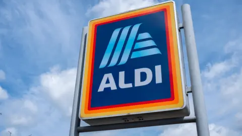 Mike Kemp/In Pictures via Getty Images A large sign for Aldi on grey poles set against a blue sky.
