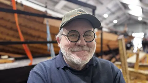 Jo Black/BBC David MacDonald stands in front of the ship reconstruction project and smiles at the camera. He wears a green cap, blue jumper and blue polo top with a collar underneath. He has circular glasses on and he has a grey beard.