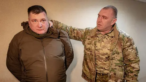 Security Service of Ukraine Ukrainian SBU chief Vasyl Malyuk (right) stands with his hand behind an arrested man's head, apparently holding him by the hood of his jacket. Malyuk is  dressed in military-style fatigues and the other man is wearing a zipped-up brown jacket.