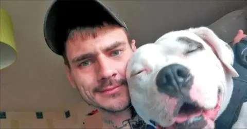 A man with a baseball cap looks into the camera. He is holding a white dog with its eyes closed
