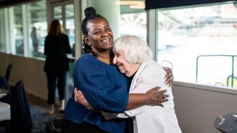 Teju Adedayo hugs Betty Brown. Ms Adedayo has dark hair styled in braids and is wearing a blue top. Ms Brown has grey hair and is wearing a white jacket.