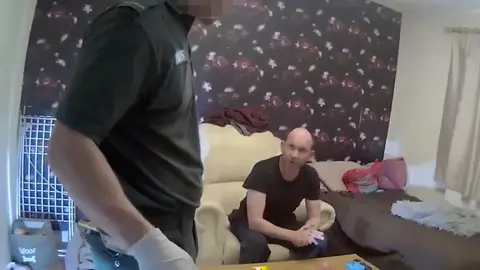 West Mercia Police Man sat down on a cream coloured chair in a living room with purple flowery wallpaper. He has his hands clasped and is wearing a black T-shirt. A paramedic is stood near him.