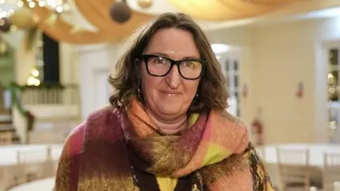 Martin Giles/BBC Karen Thomas smiles at the camera inside a hall. She has shoulder length brown hair, wears glasses and a red and yellow chequered scarf. 