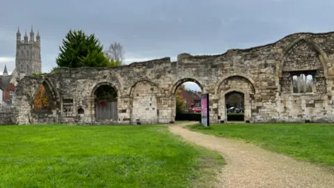 BBC The remains of a medieval wall, with several brick arches and the turret of Gloucester Cathedral visible in the background. There is an information board in the centre on a large grassy area in front of the wall