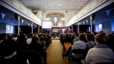 A crowd of seated people watching three musicians perform on stage. There are two balconies either side of the photo leading to the front, with fairy lights strung between them