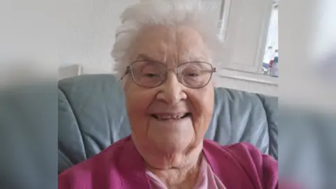 An elderly woman, seated in an armchair, smiles at the camera. She has white hair and glasses and is wearing a light pink blouse and a darker pink cardigan.
