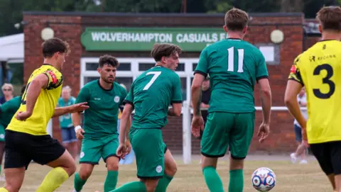 Three footballers in green shirts and two in yellow shirts are on a pitch with little green grass at this angle. A building in the background includes the words Wolverhampton Casuals on a sign.