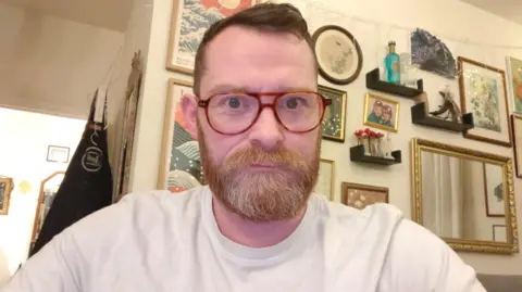 Alex Young in a light brown T-shirt, sitting looking directly at the camera with a wall behind covered in picture frames