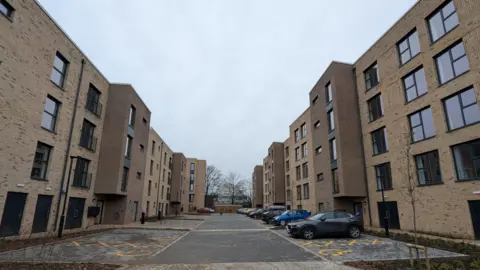 Two blocks of flats sit either side of a pedestrianised road. The flats are brown in colour and a row of cars are parked to one side.