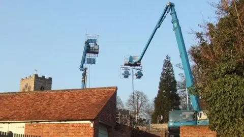 Carol Sawbridge A view of a crane during the filming of Les Miserables in Ewelme. It towers above the buildings. It is a sunny day.