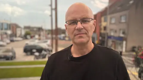 A man wearing a black t-shirt stands in front of a row of shops , outside of which a number of cars are parked. He is wearing eye glasses and is clean shaven and bald headed.