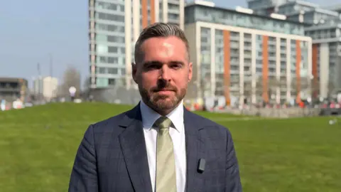 Tom Copley in a dark grey chequered suit, gold tie, and white shirt looks into the camera. He is stood in front of some multi-storey buildings in front of a lawn of grass. 
