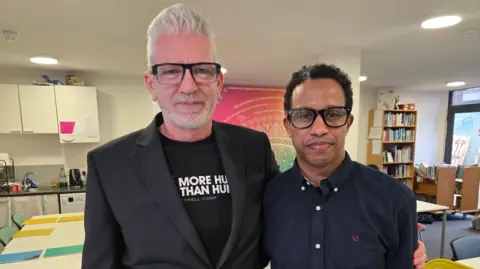 Simon and Ilyass stand arm in arm. Both are wearing dark clothing, and have black glasses on, and are seen in a classroom setting with bookcases in the background