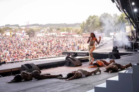 Ayra Starr performs on Glastonbury's Pyramid stage. Thousands of fans can be seen at the foot of the stage, while the musician's dancers lie protsrate on the floor.