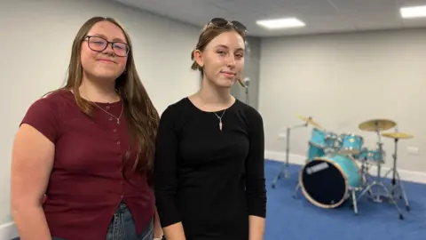 BBC Lara has long loose brown hair and glasses and is wearing a burgundy waistcoat and jeans. Isabella has her light brown hair tied up and glasses on her head. She is wearing a black top. There is a blue carpet and a drum kit in the background