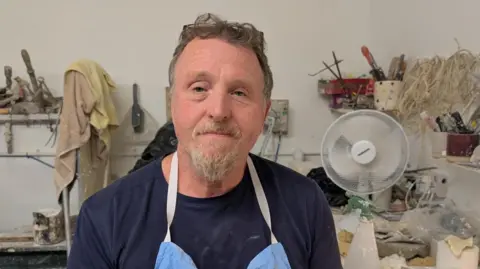 Andrew Henshall wears an apron, he has a beard and is smiling at the camera. He sits in front of his workbench, where there is a fan and plenty of tools and materials.
