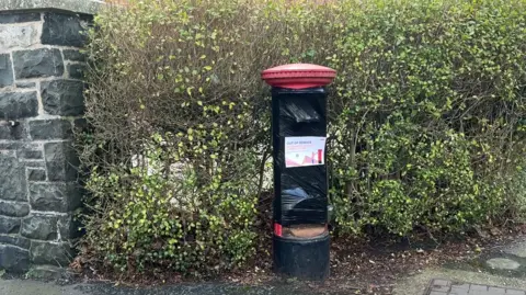 Post box with red top wrapped in black tape and marked with an “Out of Service” sign stands beside a stone wall and a trimmed hedge on a residential street.