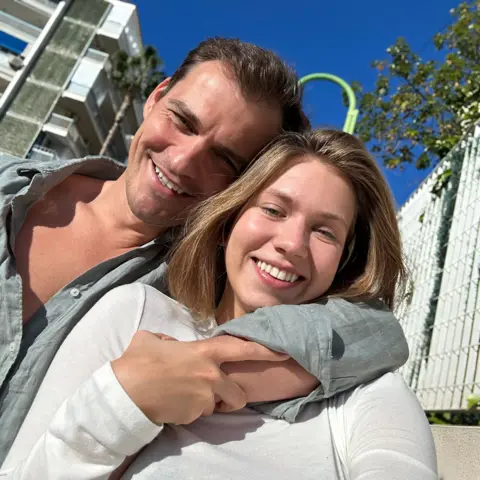 Handout Two people standing close together in the foreground, with one arm wrapped around the other’s shoulders. Both are wearing light long‑sleeved tops. The scene is outdoors in bright sunlight with a block of modern apartments behind them.
