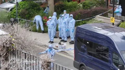 Jonathan Brady/PA Media A group of people wearing forensics suits. The suits are blue and the people are wearing surgical face masks and latex gloves. Police tape can be seen too.