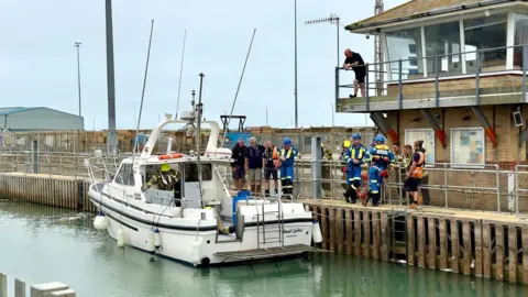 A white yacht sits in Shoreham Harbour, with emergency teams wearing blue and yellow standing on the boat and harbour, and two civilians standing on the yacht's deck.