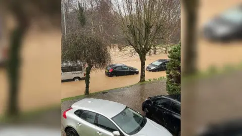 Tracey Trotter A flooded road seen from the upper window of a house, with cars submerged up the upper part of their wheels.