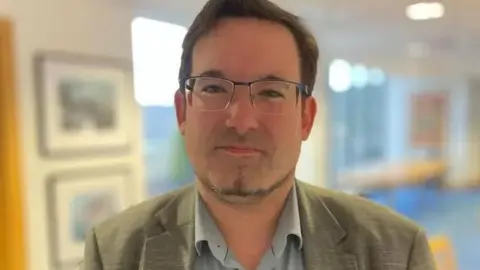 Lawrie Hooper, a man with brown hair and glasses, he stands in a corridor.
