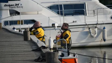 Shaun Whitmore/BBC Two emergency services workers, wearing yellow buoyancy suits, stand in an orange boat which is moored at a quayside. A larger white pleasure craft can be seen behind them.