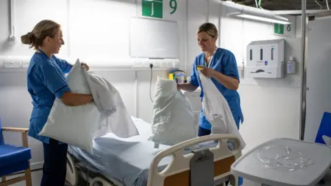 PA Media Two female nurses, both with blonde hair wearing blue scrubs, stand either side of a bed in a brightly-lit medical ward. They are putting pillow cases on pillows.