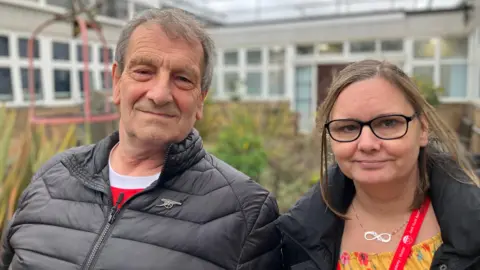 Shariqua Ahmed/BBC Ian, a man wearing a black zipped jacket and Sam, a woman wearing a yellow top and a black coat, smiling for camera.