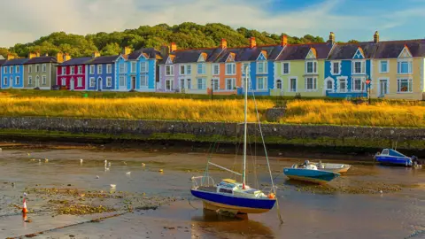Getty Images A view from the harbour looking at a row of colourful houses in Aberaeron. Each town house is a different colour. A bank of grass between the houses and the water catches the light, with boats anchored up - the tide has gone out. 