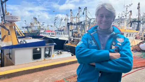 Ian Perkes is smiling with his arms crossed as he stands in front of fishing boats in Brixham on a sunny day. He has white hair and is wearing a bright blue sailing-style anorak which has badges on it.