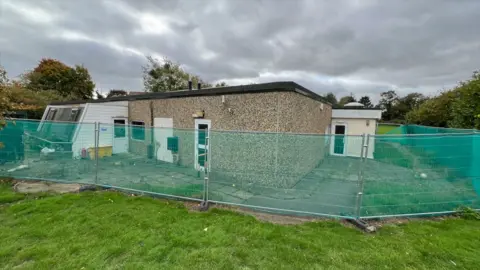 Stuart Woodward/BBC A single storey school building with a flat roof and pebbledash walls, with two white coloured extensions at either end. The building is surrounded by metal fencing, and there is grass in front of the building with grey skies above