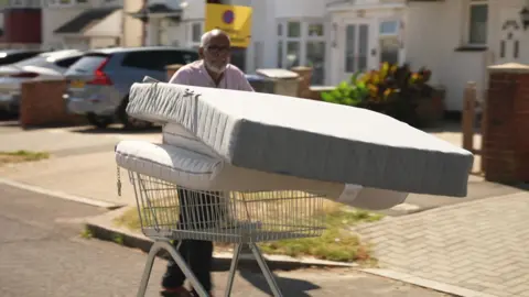 An older man pushes a shopping trolley loaded with mattresses along a residential street.