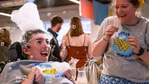 A man sits in a wheelchair. He wears a grey sweatshirt which has the words 'wonder bubbles' on it, in yellow writing. The man has a collection of small bubbles on his head. A woman stands to his left, she wears the same top, but with a blue skirt. She is holding a bubble blower. 