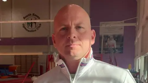 James Patient/BBC Scott Hann has a shaved head and is wearing a white Team GB jacket with tall collars. He is standing on the floor of a gymnastics club, with a pole behind him.