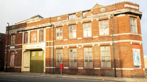 Sunderland City Council The two-storey red-bricked building has large green doors with the letters "P" and "S" painted on them. There are several large boarded-up windows.