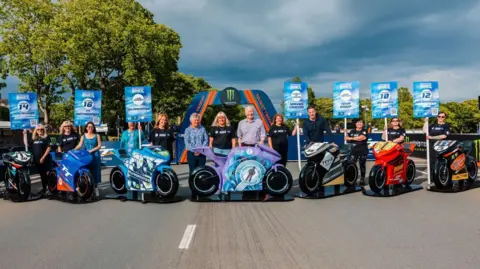 Seven of the colourful bike models are lined up on a closed road, which is the start of the TT course. People stand behind them, some hold signs up explaining the designs. You can see large green trees in the background.