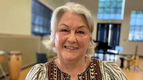 Alex Dunlop/BBC Chris Wiley has short white hair. She is smiling and is wearing a patterned white and brown T-shirt and a silver necklace. She is sitting in a large room which has several windows and a fire exit. 