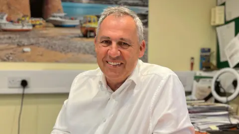 Andy is in a white shirt sits behind a desk in an office. The desk contains an open notebook, computer keyboard, mouse, and paperwork. Behind the desk are noticeboards, office equipment, and a large wall photograph showing a coastal harbour scene.