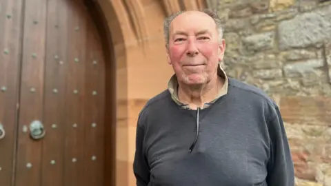 A man with receding hair and wearing a dark blue top stands in front of a church