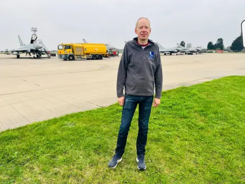CAA A man standing on grass in front of a fighter jet on an airfield.