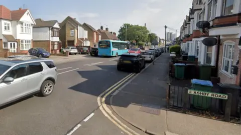 Ashby Road, but Gammons Lane, Watford. There is a road sign, cars parked, a cars driving down the road and a blue bus in the distance. You can see two rows of houses along a road, that are mainly brick, with two satellite dishes. 