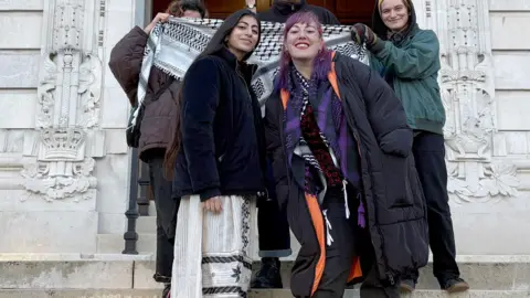 Press Association Hiba Ahmed and Ayeshah Behit smile for the camera on the steps outside Cardiff Crown Court. Three people behind them hold up a black and white Palestinian-style scarf. Hiba Ahmed has long dark brown hair and is wearing a blue coat with a white floor length, white skirt. Ayeshah Behit has pink/purple hair and is wearing a long black puffa coat, with black trousers and a purple Palestinian-style scarf around her neck. 