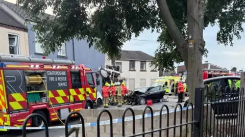 Jason Powner Firefighters can be seen gathered around a building, with a damaged car to the side of them. There are two fire engines and an ambulance on the road
